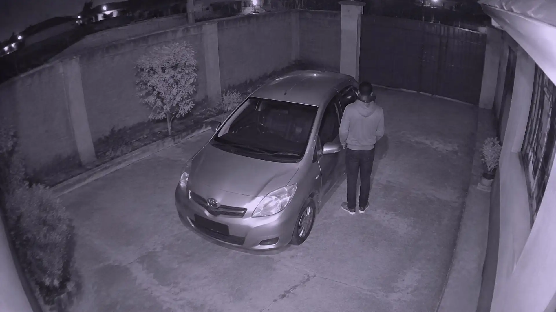 Night-vision camera view of a car parked in a driveway, with a person standing close to it.