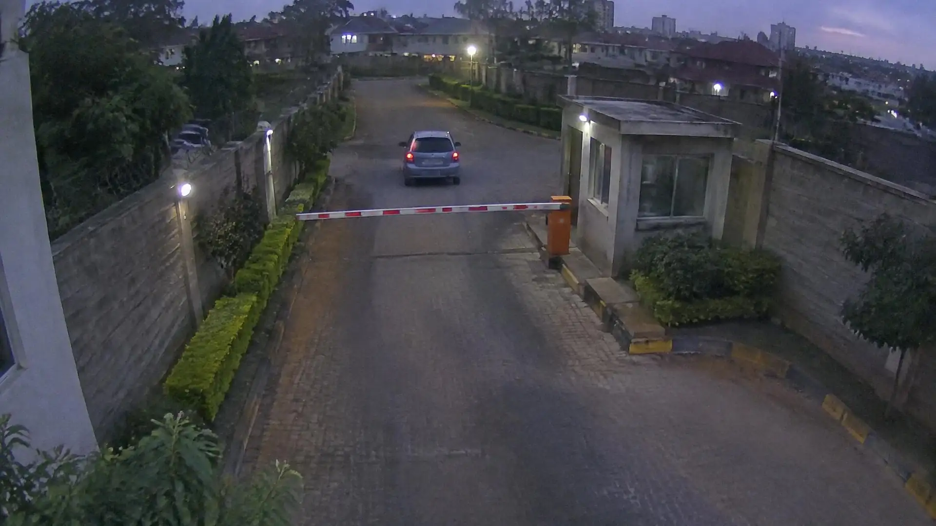 Camera view of an estate entrance at dusk with a boom barrier, guard booth and an approaching car.