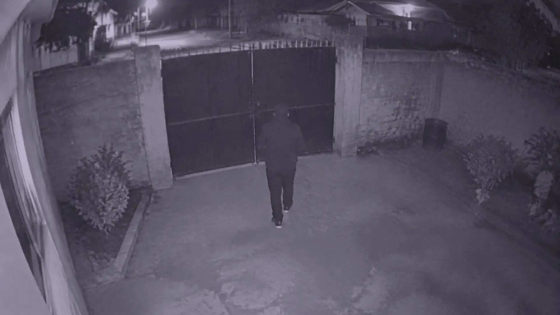 Night-vision camera view of a steel gate in a perimeter wall, with a person approaching it.