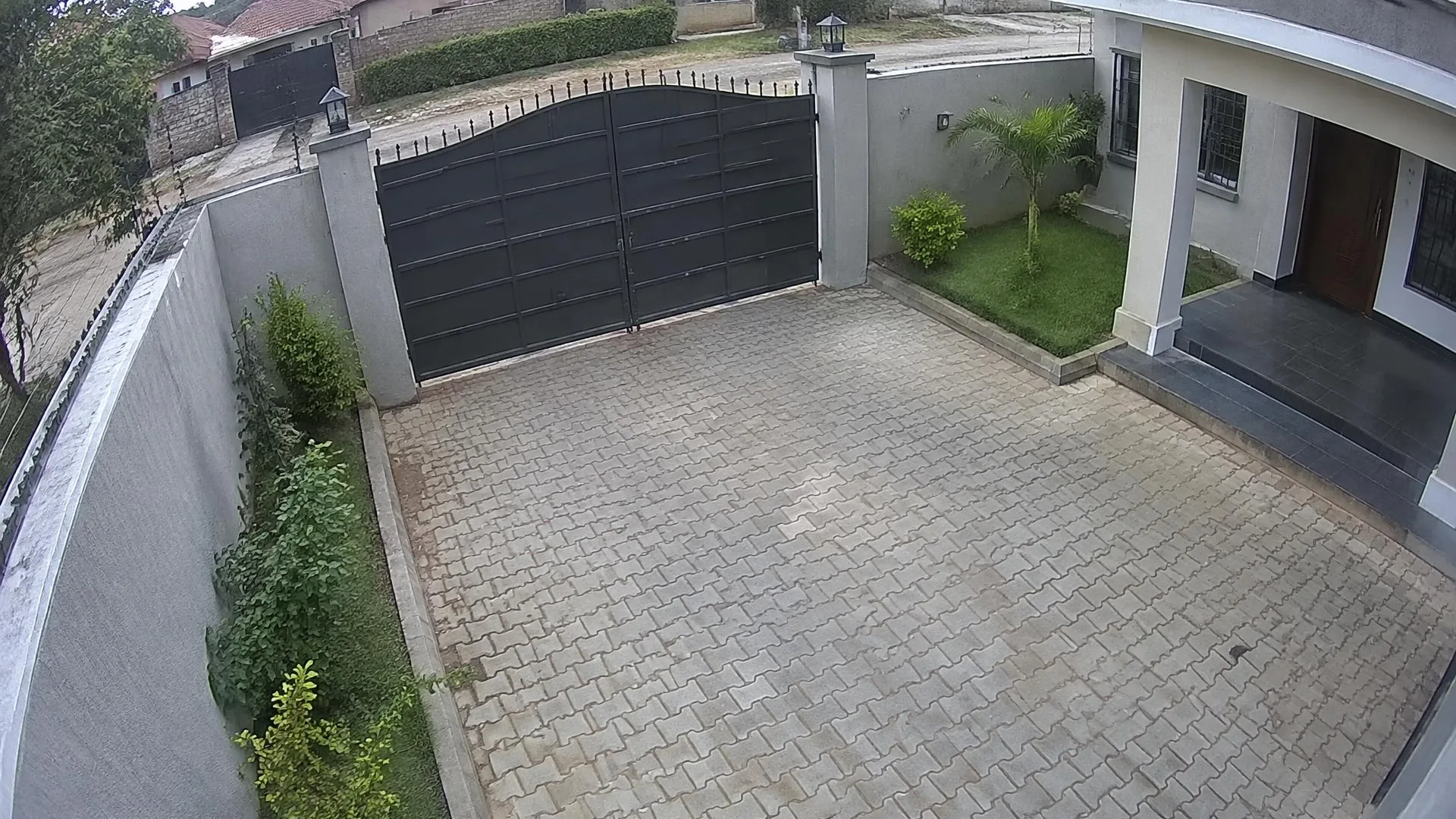 High wall camera view of a Nairobi home compound with a steel gate, paved courtyard and house entrance in daylight.