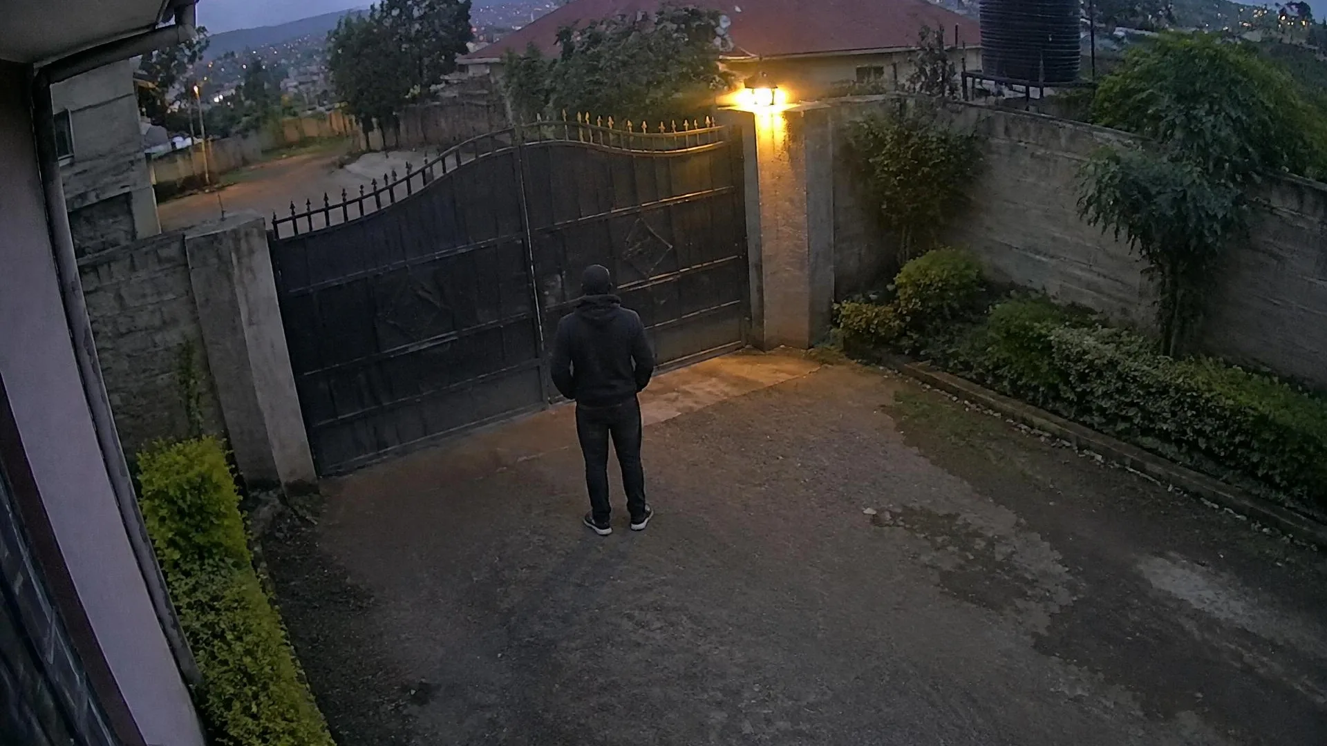 Dusk camera view of a person standing still outside a residential steel gate, seen from behind.