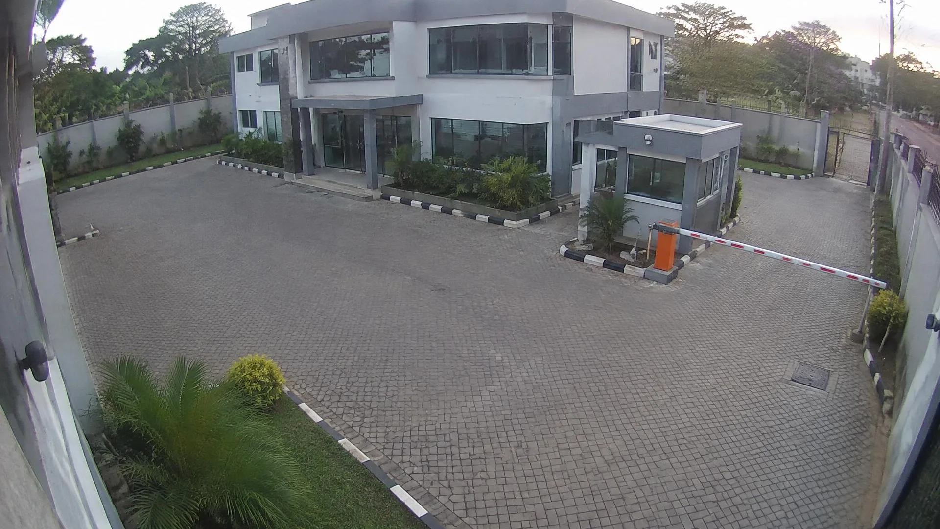 Camera view of a Nairobi office courtyard with a two-storey building, guard booth and boom barrier in late afternoon.