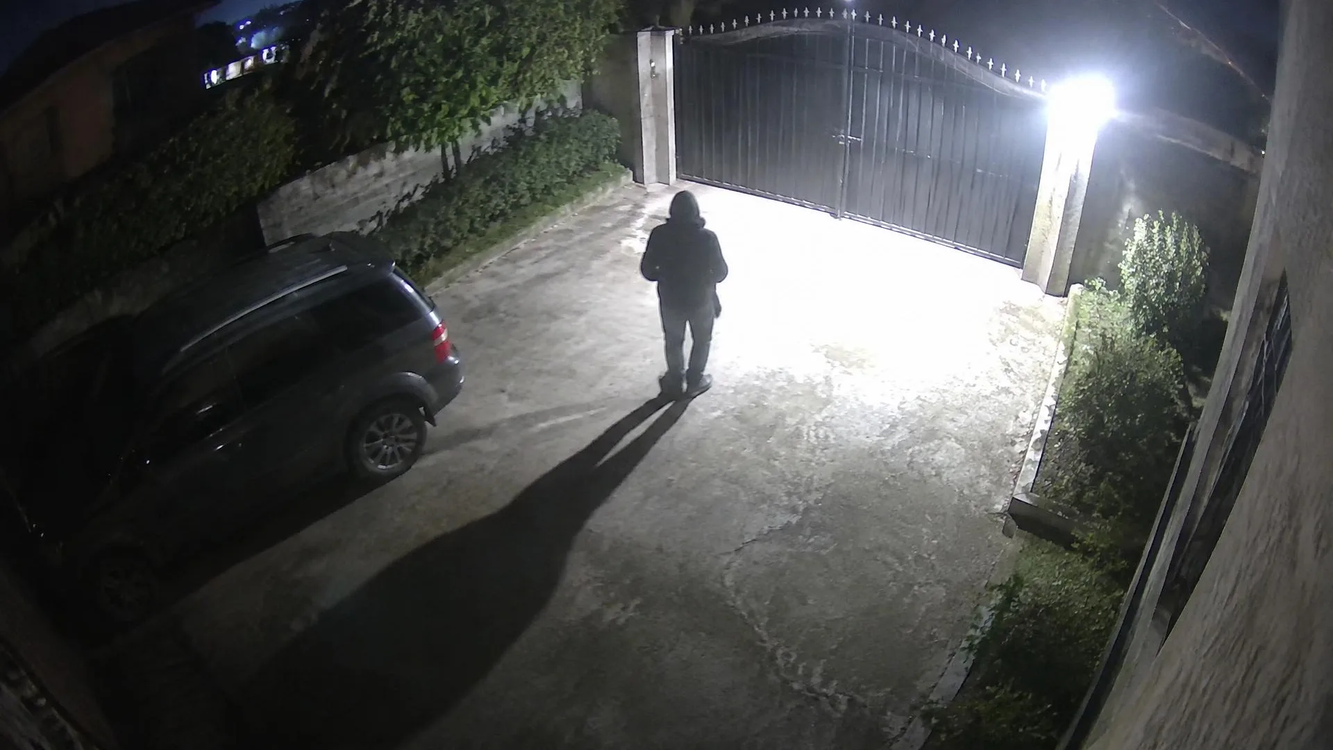 Night camera view of a driveway and gate brightly lit by a security spotlight, with a person silhouetted in the glare.
